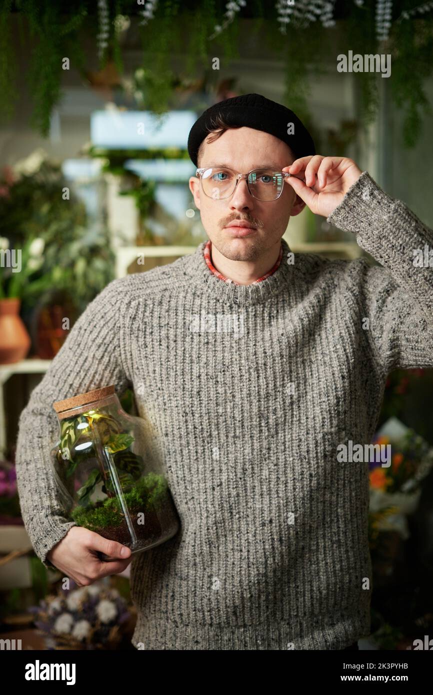 Joli barbu mâle tenant le pot avec plante dans le magasin de fleur. Bon homme floriste qui porte un chandail, un bonnet et des lunettes. Concept de petite entreprise. Image verticale de haute qualité Banque D'Images