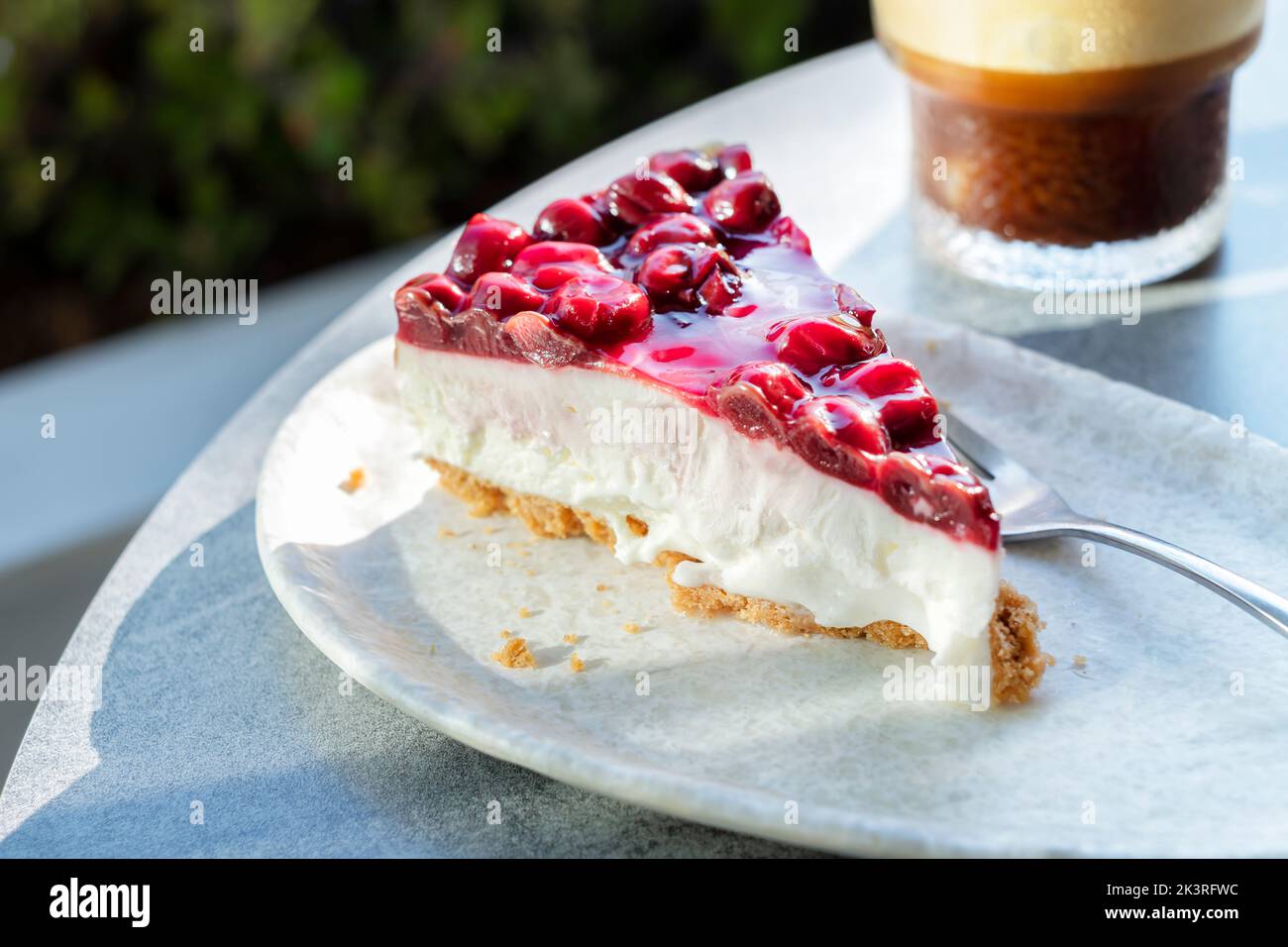 Une seule tranche, ou portion, de gâteau au fromage de cerise fraîchement préparé. Il est servi sur une assiette avec un café glacé. À une table extérieure dans un café. Banque D'Images