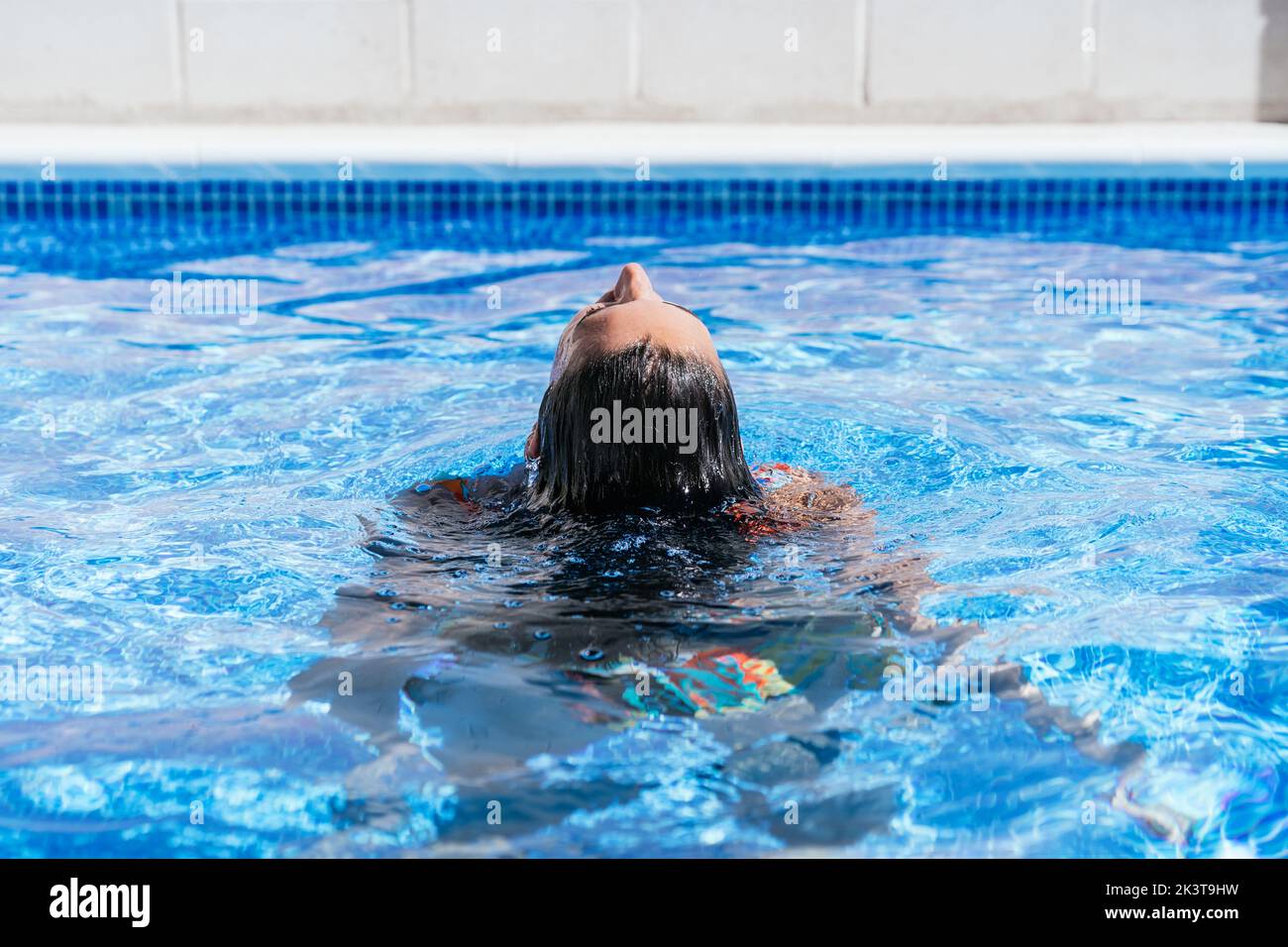 Vue arrière de la jeune femme détendue dans le bikini coloré flottant avec les yeux fermés dans l'eau de cristal de la piscine bleue tout en appréciant les vacances d'été Banque D'Images
