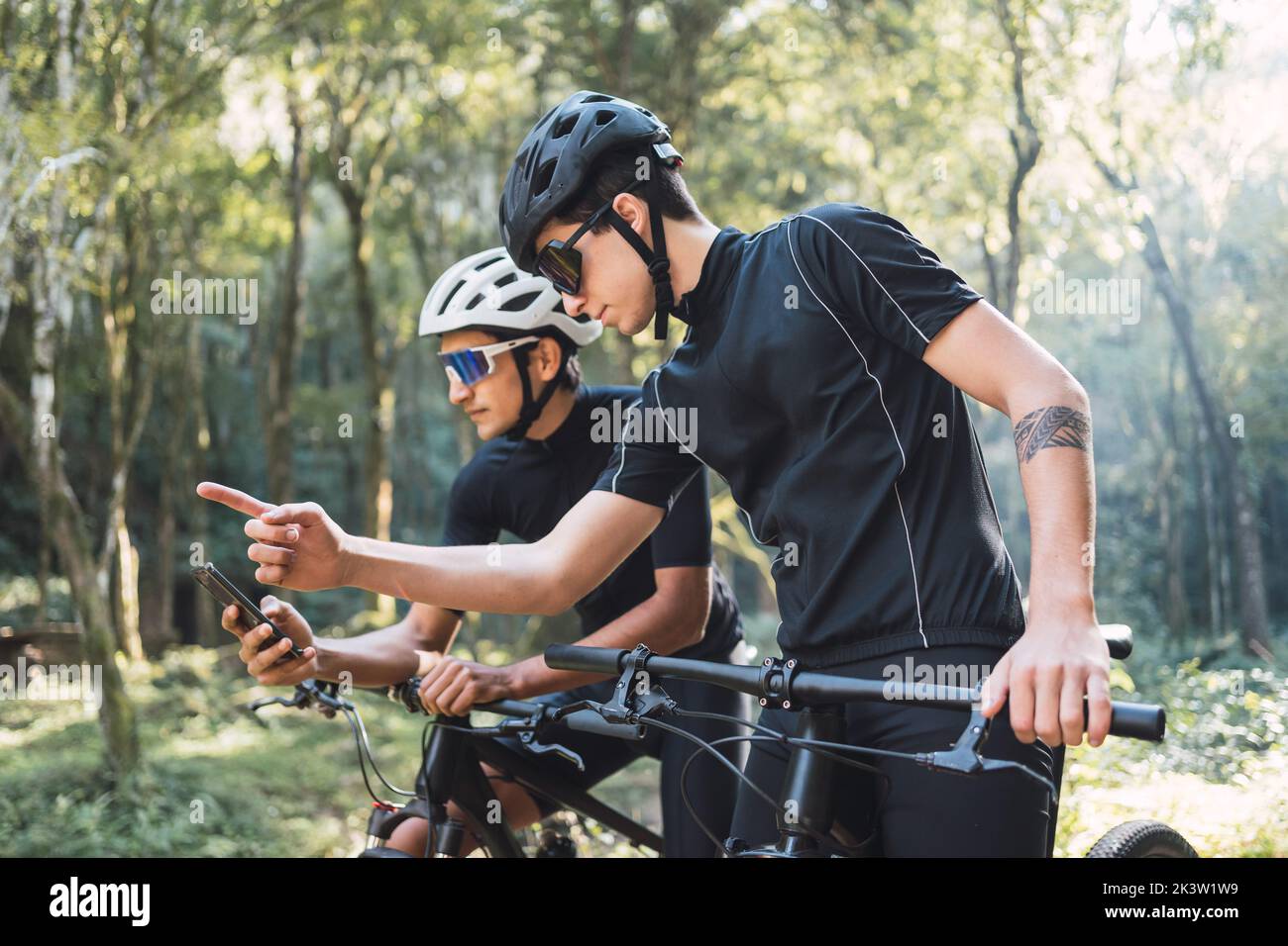 Jeune cycliste masculin tatoué pointant du doigt vers l'extérieur, montrant la direction tout en vérifiant l'application GPS sur le téléphone portable pendant un voyage à vélo avec un ami dans les fores Banque D'Images