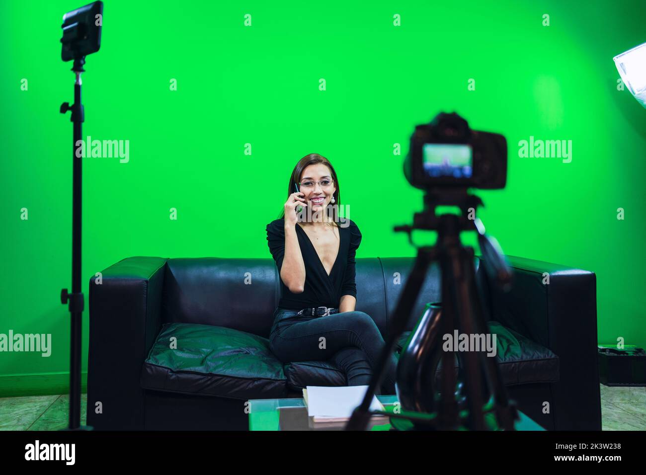 Influenceur positif souriant largement pendant un appel téléphonique tout en étant assis sur un canapé contre un mur de chromakey et en filant de la vidéo Banque D'Images