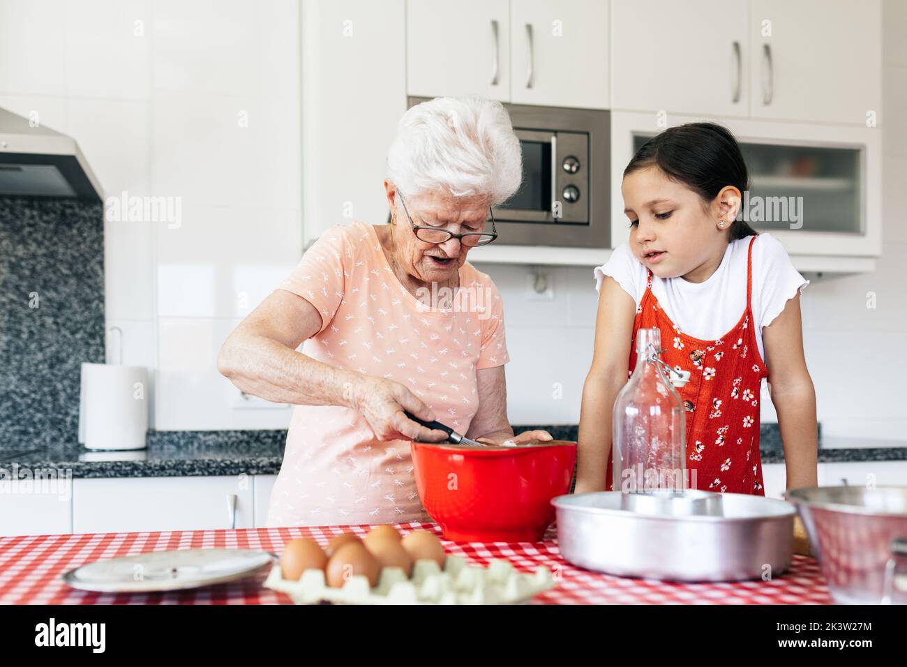Grand-mère en lunettes avec fouet près de la petite-fille cuisant de la pâte à la table dans la maison Banque D'Images