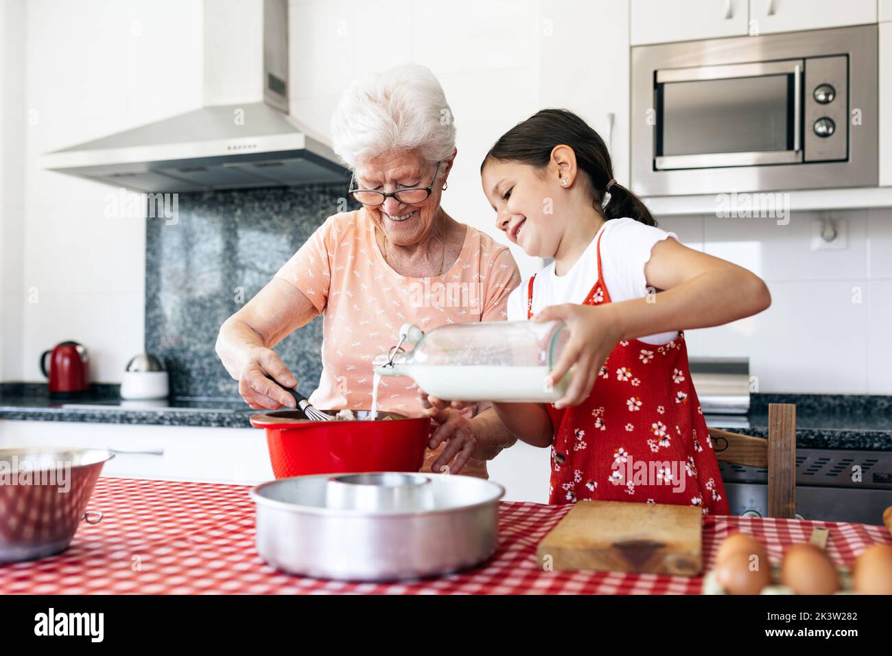 Grand-mère en lunettes avec fouet près de la petite-fille avec une bouteille de pâte de cuisson au lait à la table de la maison Banque D'Images