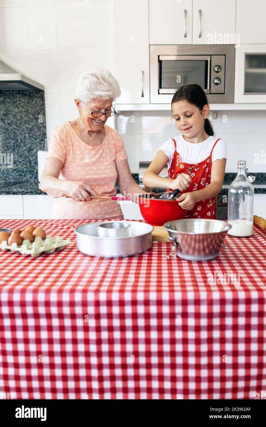 Petite-fille avec fouet près de la grand-mère dans la pâte de cuisson de lunettes à la table dans la maison Banque D'Images