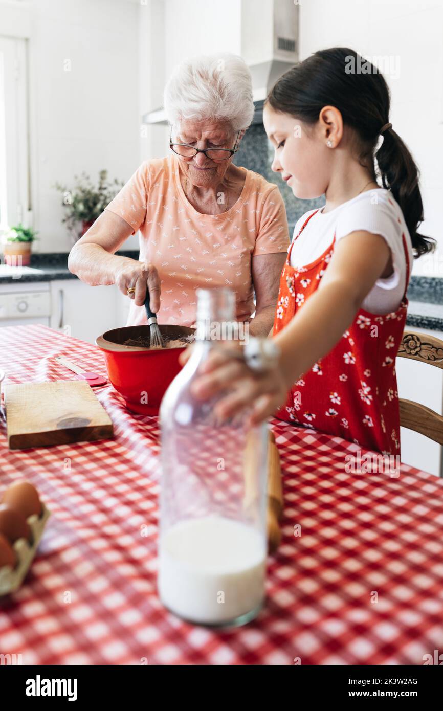 Grand-mère en lunettes avec fouet près de la petite-fille avec une bouteille de pâte de cuisson au lait à la table de la maison Banque D'Images