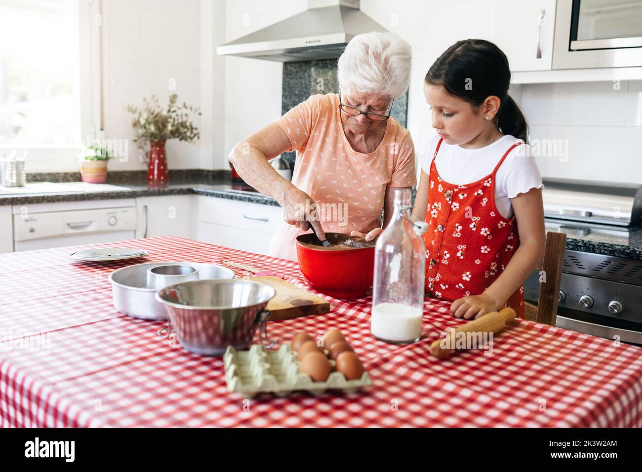 Grand-mère en lunettes avec fouet près de la petite-fille cuisant de la pâte à la table dans la maison Banque D'Images