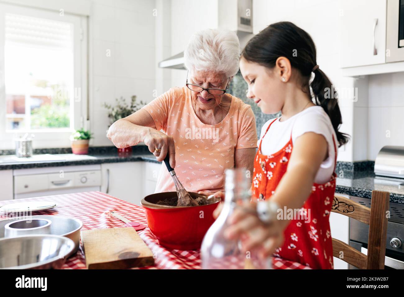 Grand-mère en lunettes avec fouet près de la petite-fille avec une bouteille de pâte de cuisson au lait à la table de la maison Banque D'Images