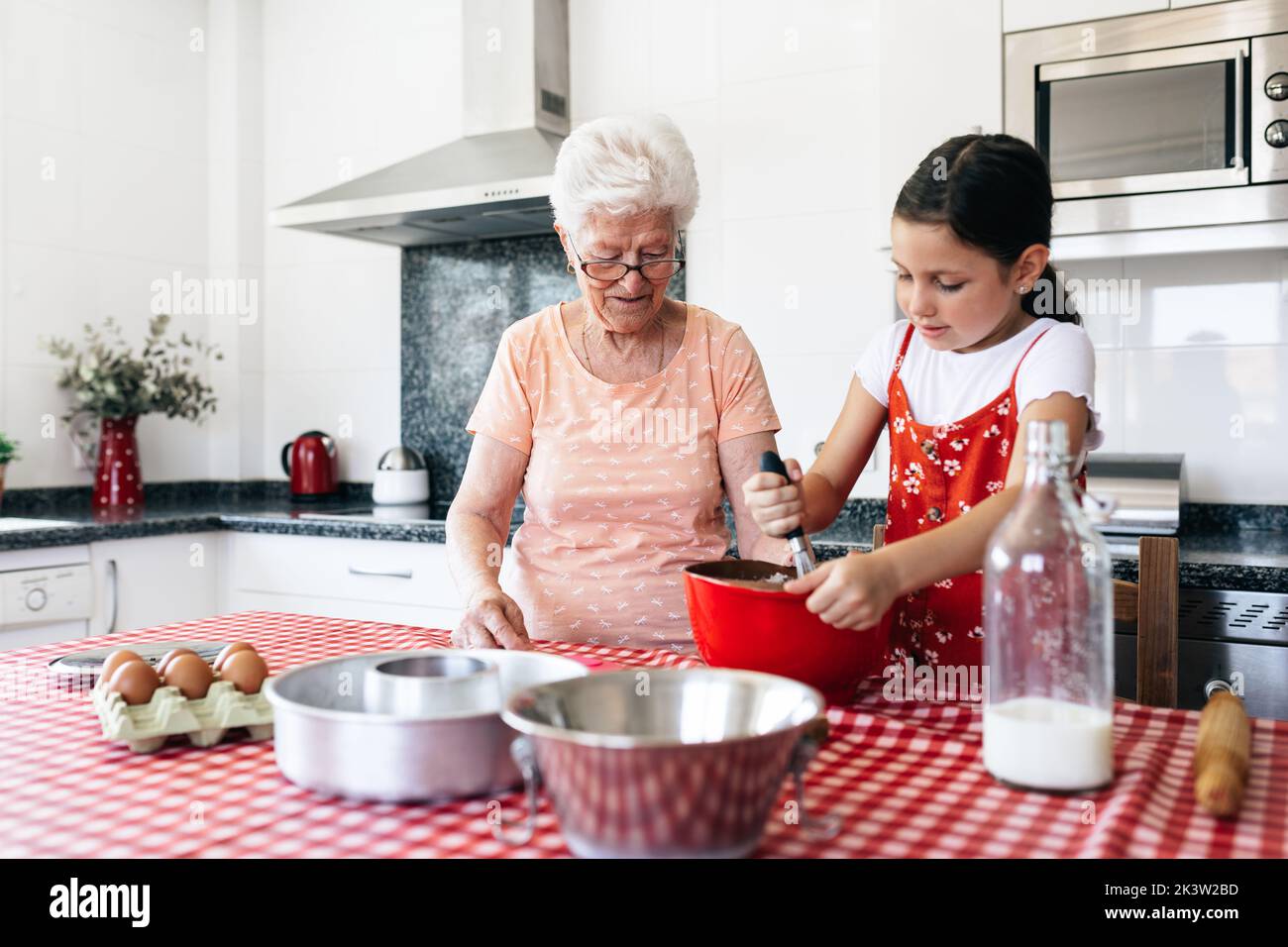 Petite-fille avec fouet près de la grand-mère dans la pâte de cuisson de lunettes à la table dans la maison Banque D'Images
