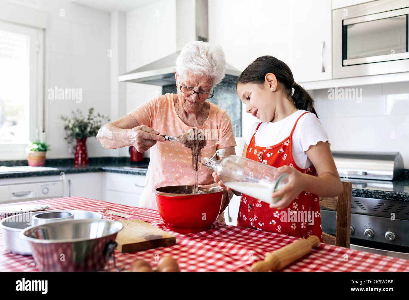 Grand-mère en lunettes avec fouet près de la petite-fille avec une bouteille de pâte de cuisson au lait à la table de la maison Banque D'Images