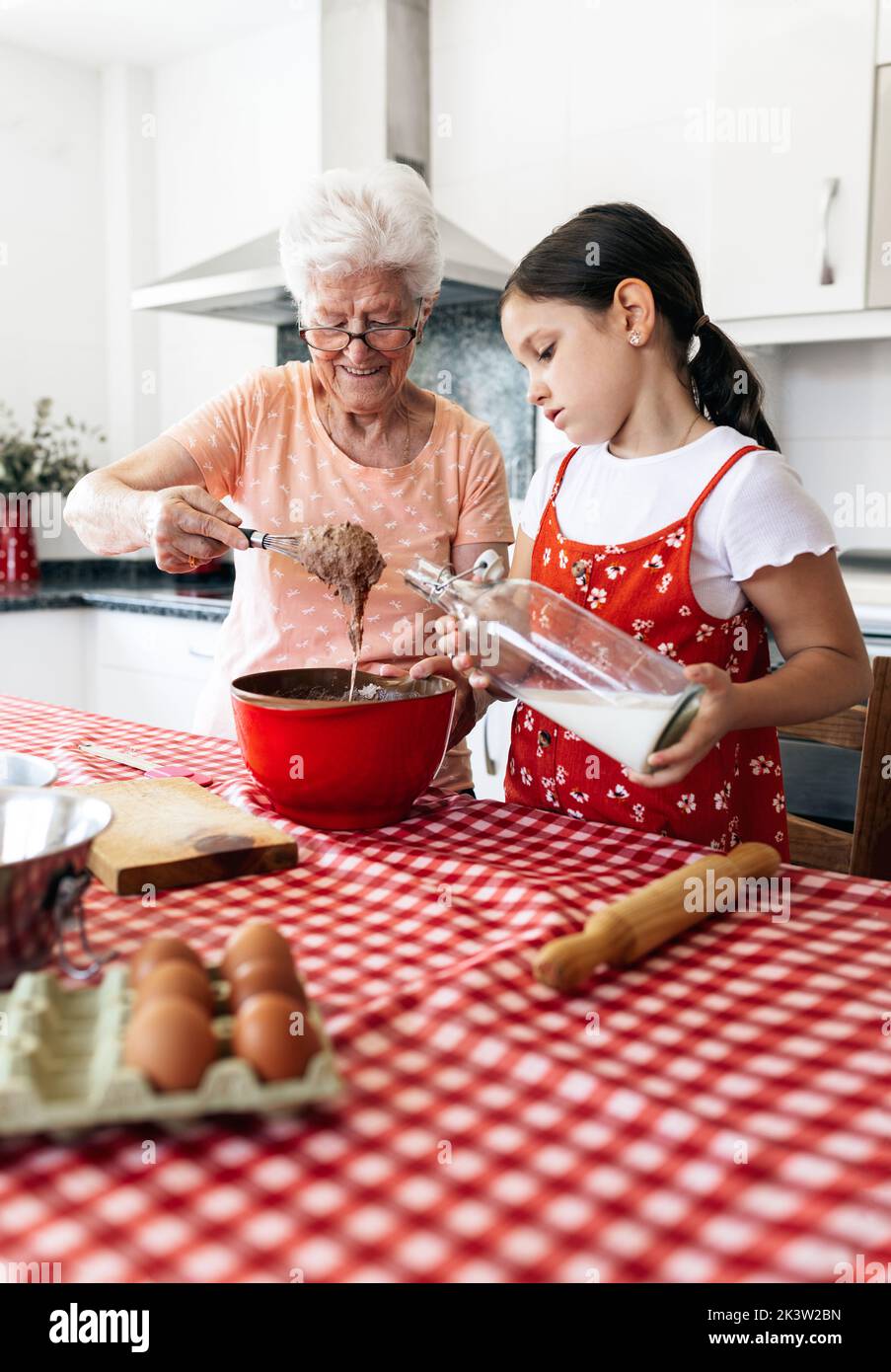 Grand-mère en lunettes avec fouet près de la petite-fille avec une bouteille de pâte de cuisson au lait à la table de la maison Banque D'Images