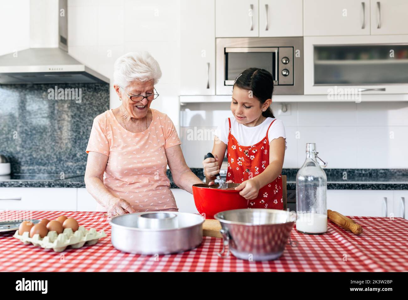 Petite-fille avec fouet près de la grand-mère dans la pâte de cuisson de lunettes à la table dans la maison Banque D'Images