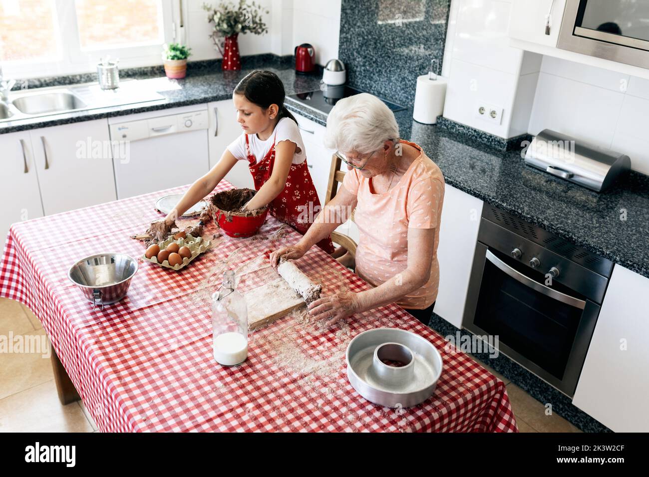 Grand-mère gaie en lunettes contre petite-fille avec de la pâte sur les mains pendant le processus de cuisson à la table dans la cuisine de la maison Banque D'Images