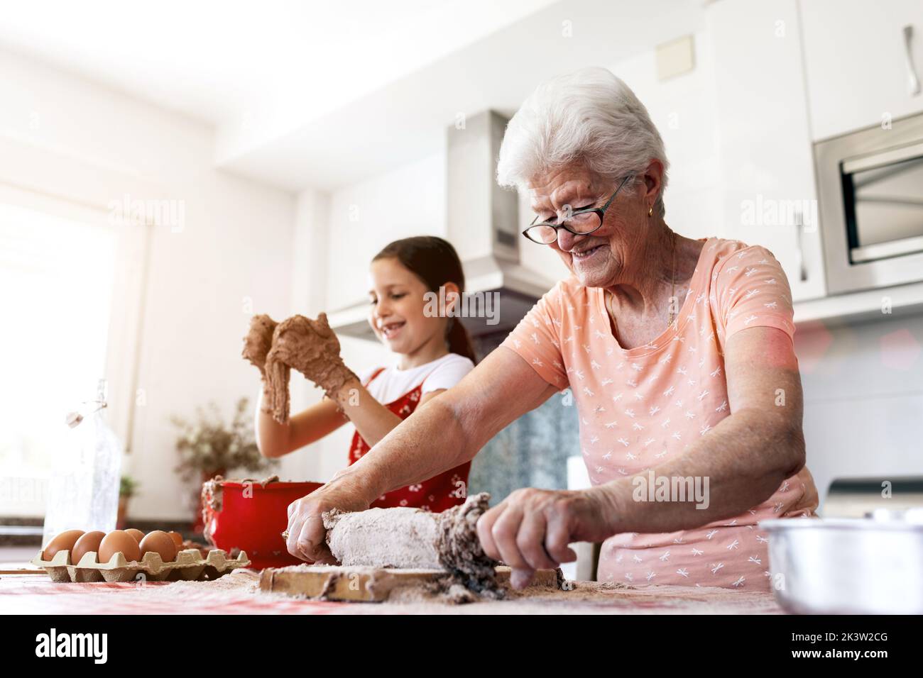 Grand-mère gaie en lunettes contre petite-fille avec de la pâte sur les mains pendant le processus de cuisson à la table dans la cuisine de la maison Banque D'Images