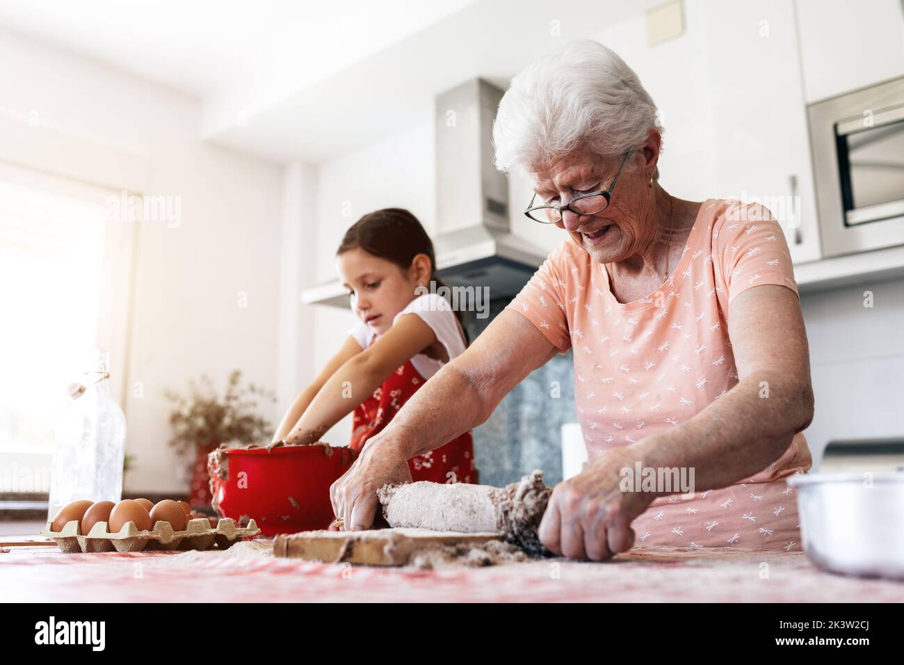 Grand-mère gaie en lunettes contre petite-fille avec de la pâte sur les mains pendant le processus de cuisson à la table dans la cuisine de la maison Banque D'Images