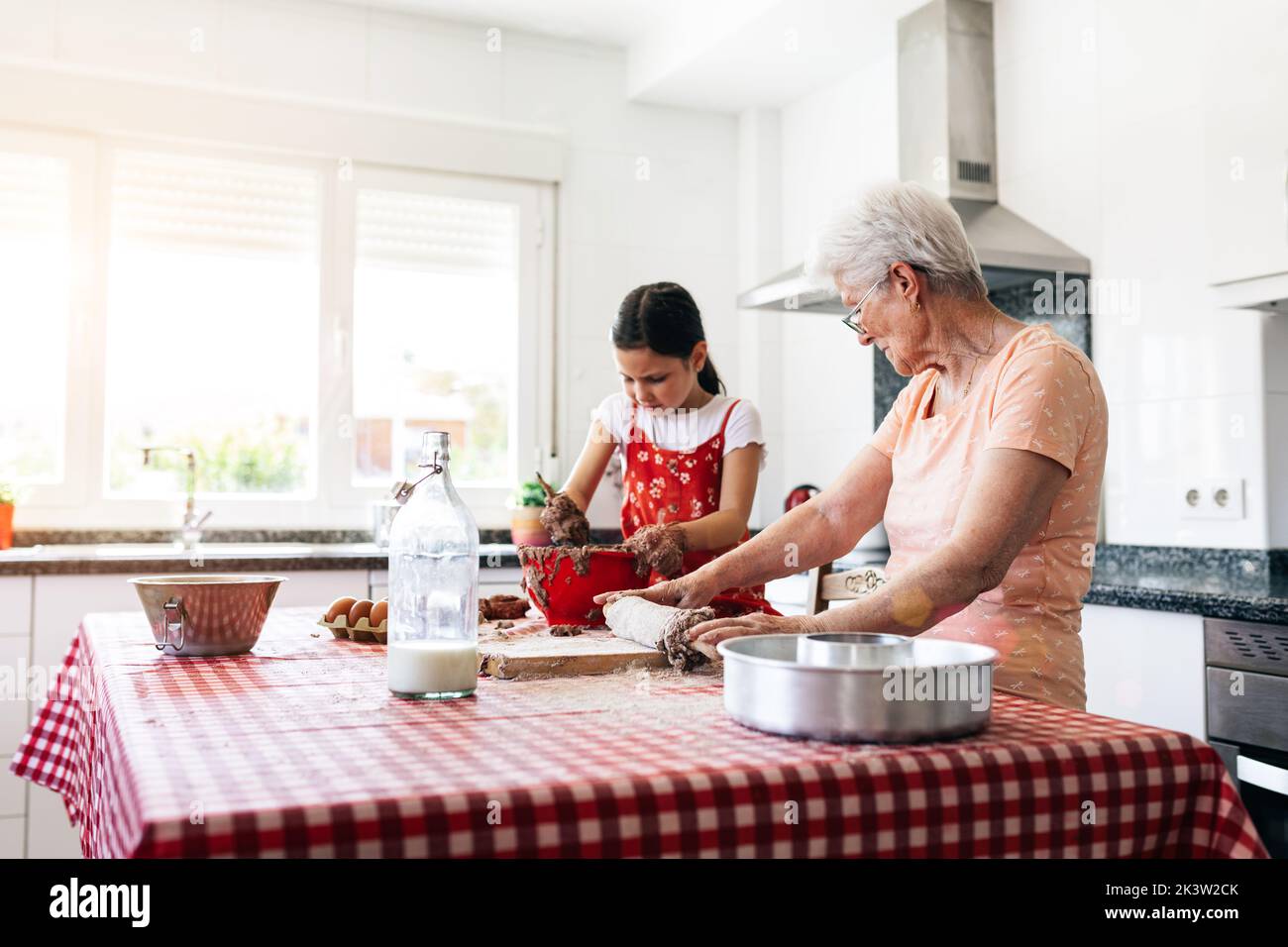 Grand-mère gaie en lunettes contre petite-fille avec de la pâte sur les mains pendant le processus de cuisson à la table dans la cuisine de la maison Banque D'Images