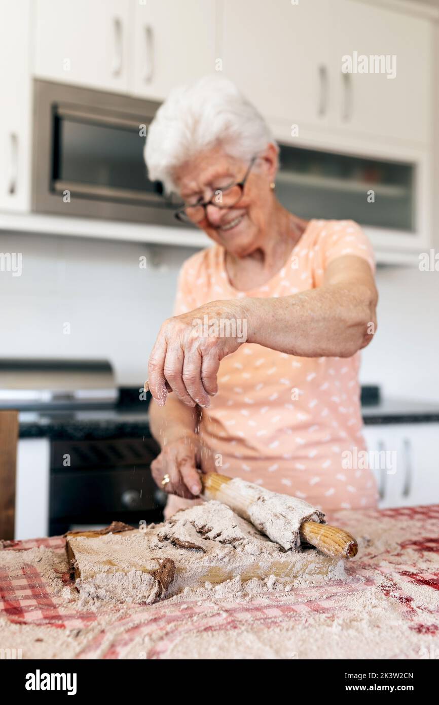 Vieille femme gaie avec des cheveux gris dans des lunettes roulant la pâte sur la planche à découper à table dans la maison Banque D'Images