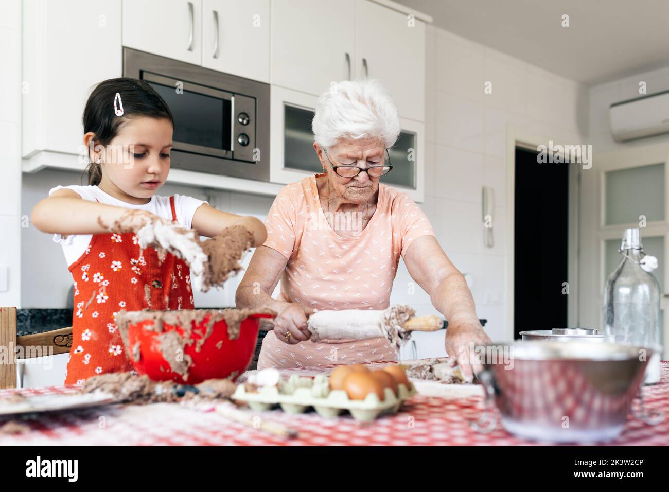 Grand-mère gaie en lunettes contre petite-fille avec de la pâte sur les mains pendant le processus de cuisson à la table dans la cuisine de la maison Banque D'Images