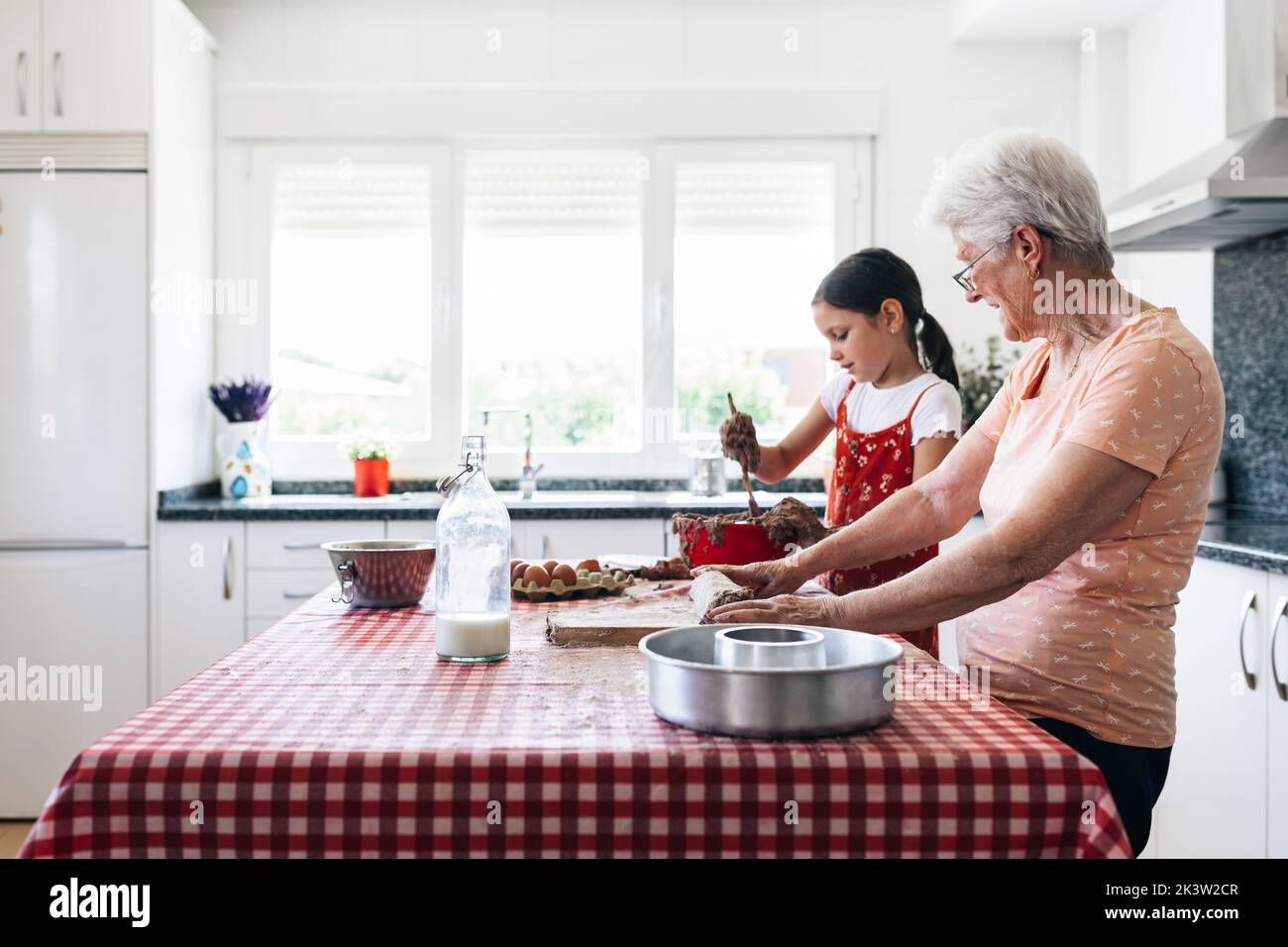 Grand-mère gaie en lunettes contre petite-fille avec de la pâte sur les mains pendant le processus de cuisson à la table dans la cuisine de la maison Banque D'Images