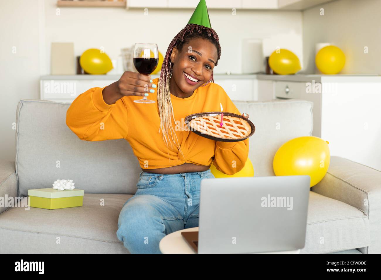 Anniversaire en ligne. Femme afro-américaine excitée célébrant le b-day, montrant une tarte et un verre de vin, les appels vidéo sur ordinateur portable Banque D'Images