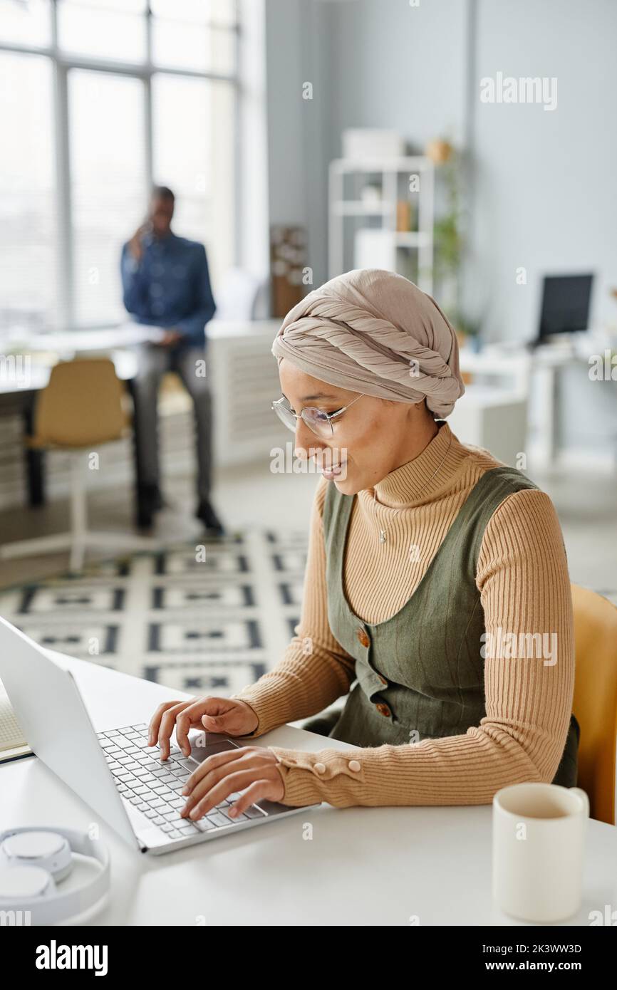 Portrait vertical d'une jeune femme d'affaires du Moyen-Orient portant un serre-tête au bureau et dactylographiant sur un ordinateur portable Banque D'Images