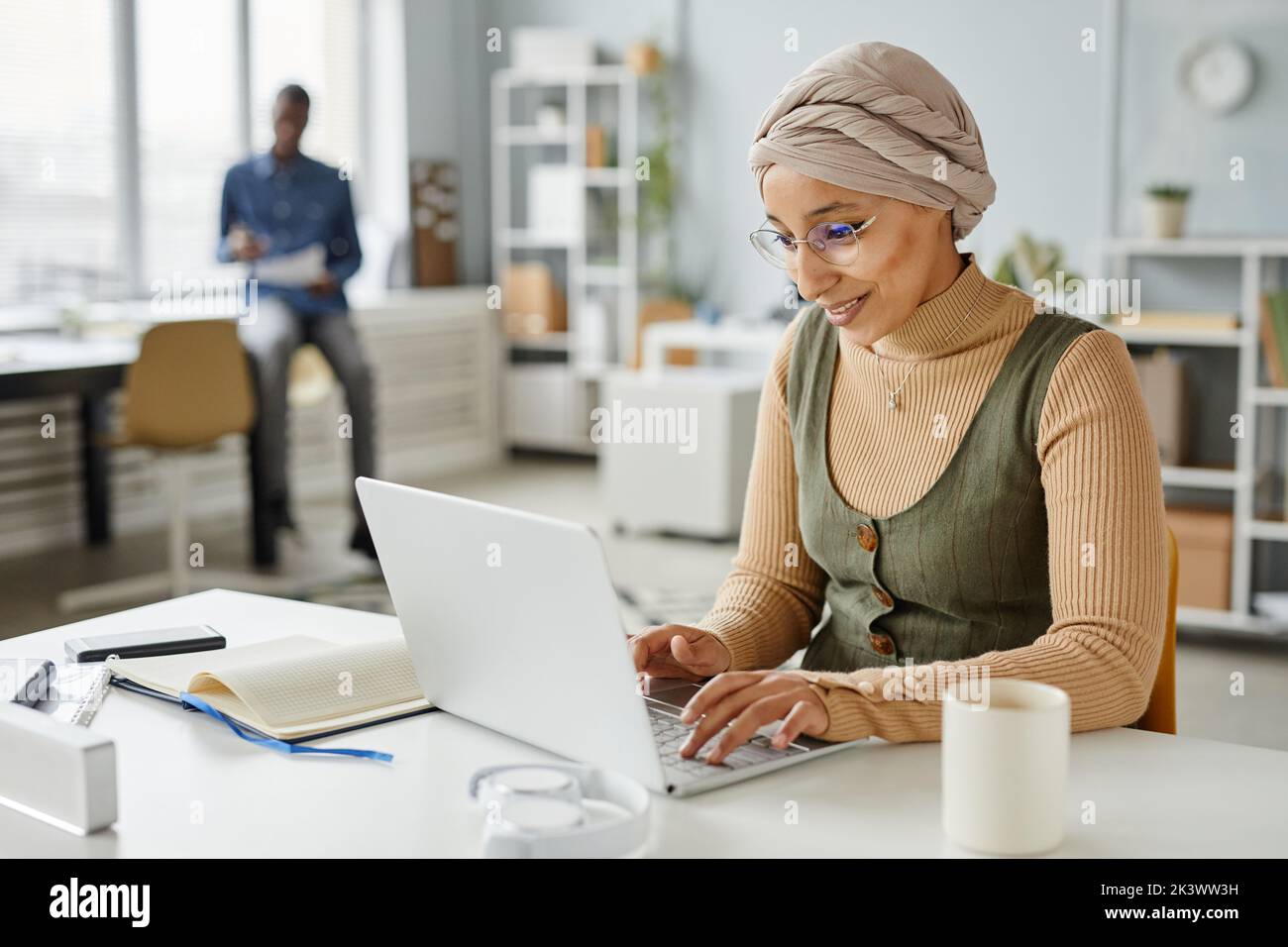 Portrait minimal d'une femme d'affaires souriante du Moyen-Orient portant un serre-tête tout en tapant sur un ordinateur portable au bureau, dans un espace de copie Banque D'Images