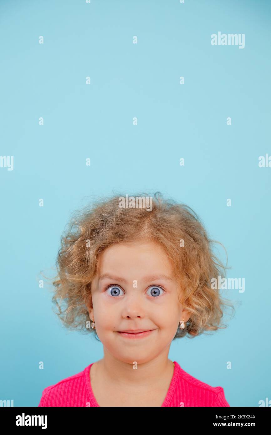 Portrait d'une petite fille souriante aux cheveux bouclés, yeux bleus larges ouverts portant un T-shirt rose, regardant sur fond bleu. Banque D'Images