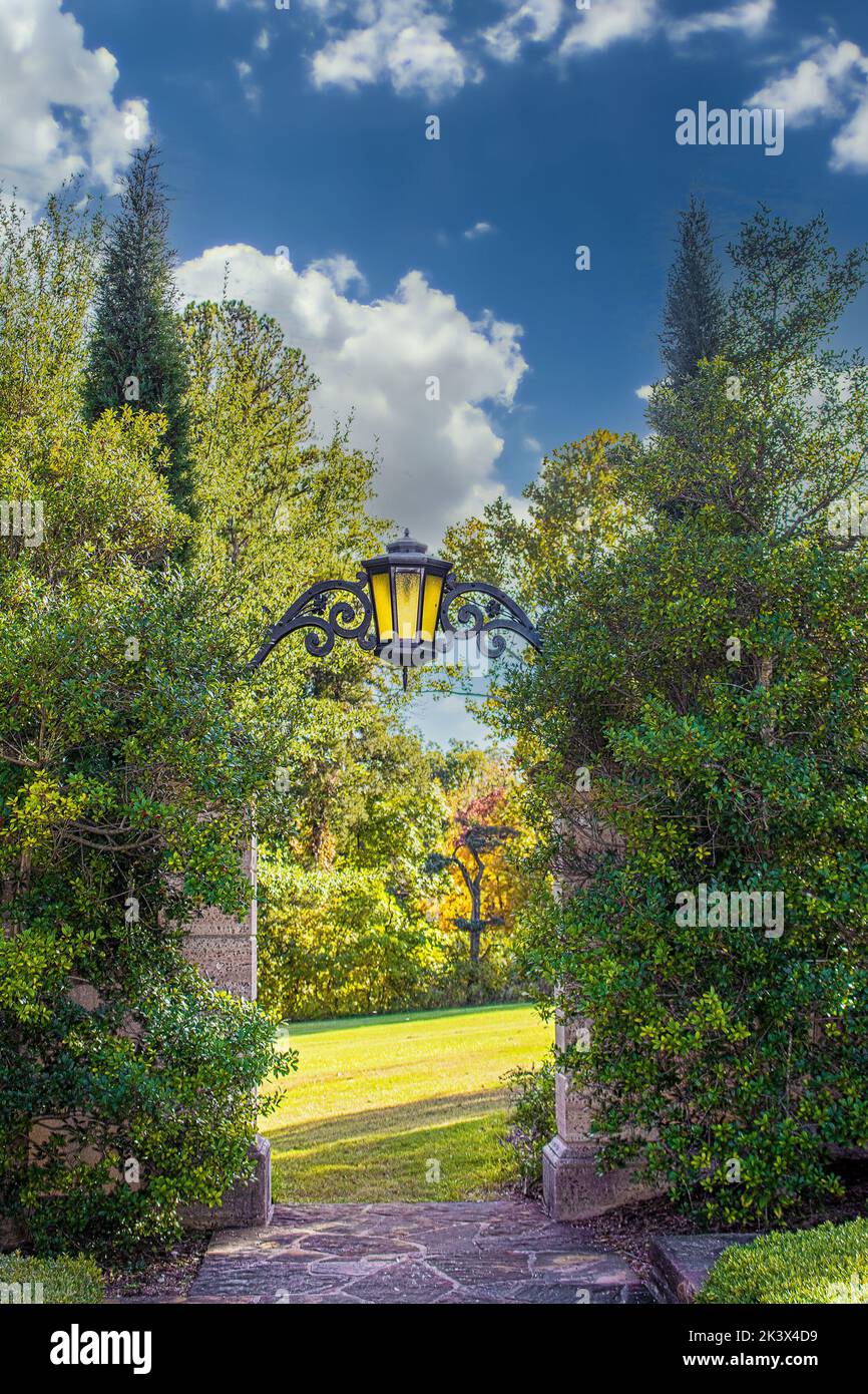 Belle porte de jardin formelle avec lanterne et beaucoup de feuillage menant à la prairie ensoleillée avec des arbres à distance Banque D'Images