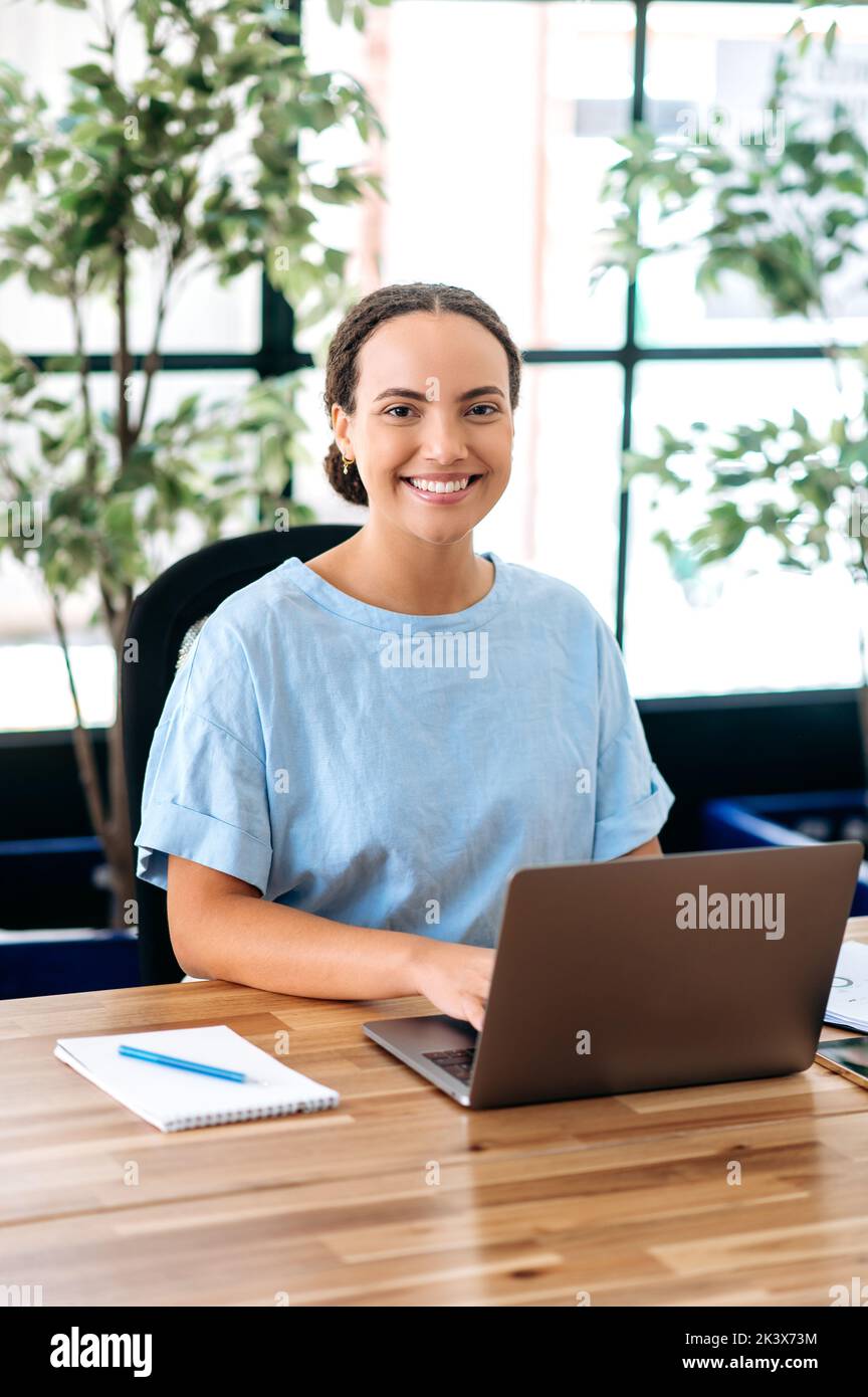 Portrait vertical d'une belle femme d'affaires. Confiante réussie latino ou brésilienne jeune femme, seo de compagnie, directeur de la création, s'assoit à un bureau dans un bureau moderne, regardant la caméra, souriant Banque D'Images