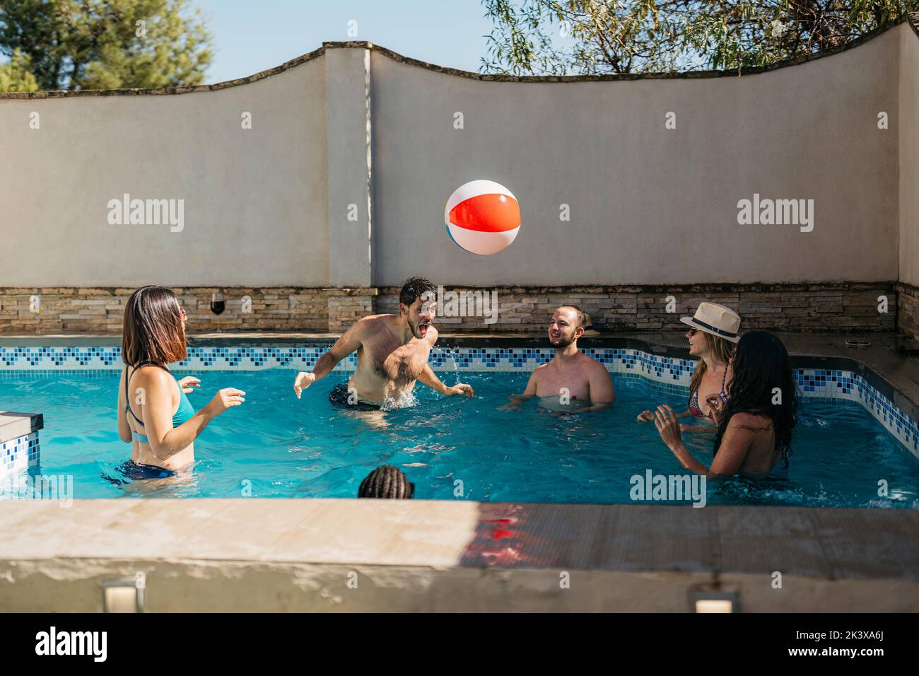 l'homme frappe une balle de plage avec sa tête dans une piscine entourée d'amis Banque D'Images