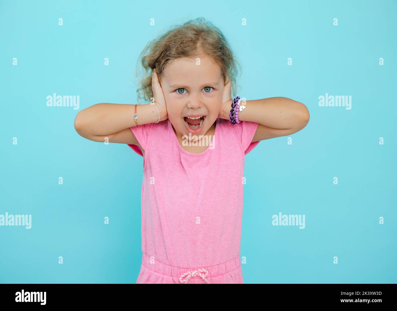 Portrait d'une petite fille à l'œil bleu qui regarde l'appareil photo, qui couvre les oreilles avec les mains, hurle sur fond bleu. Banque D'Images