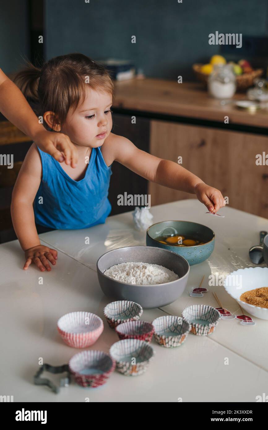 Une mère et sa petite fille cuisent en cuisine, sur une table en bois pleine d'ingrédients et de formes de cuisson. Préparer des crêpes pour le petit déjeuner à la maison Banque D'Images