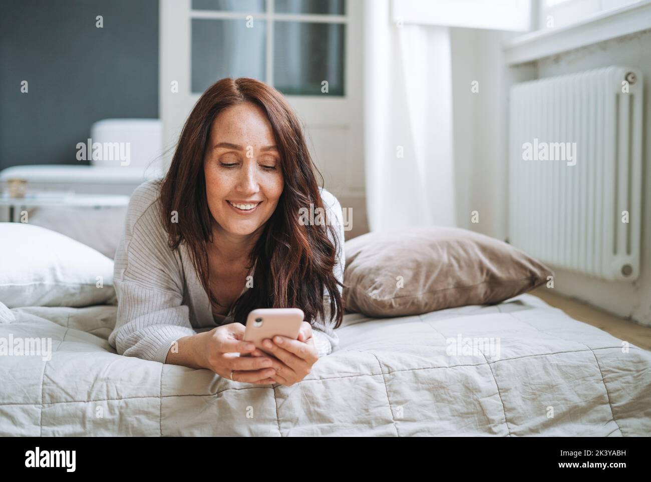 Jeune femme quarante ans avec brunette cheveux longs dans un gilet tricoté confortable utilisant un téléphone portable dans le lit à la maison Banque D'Images
