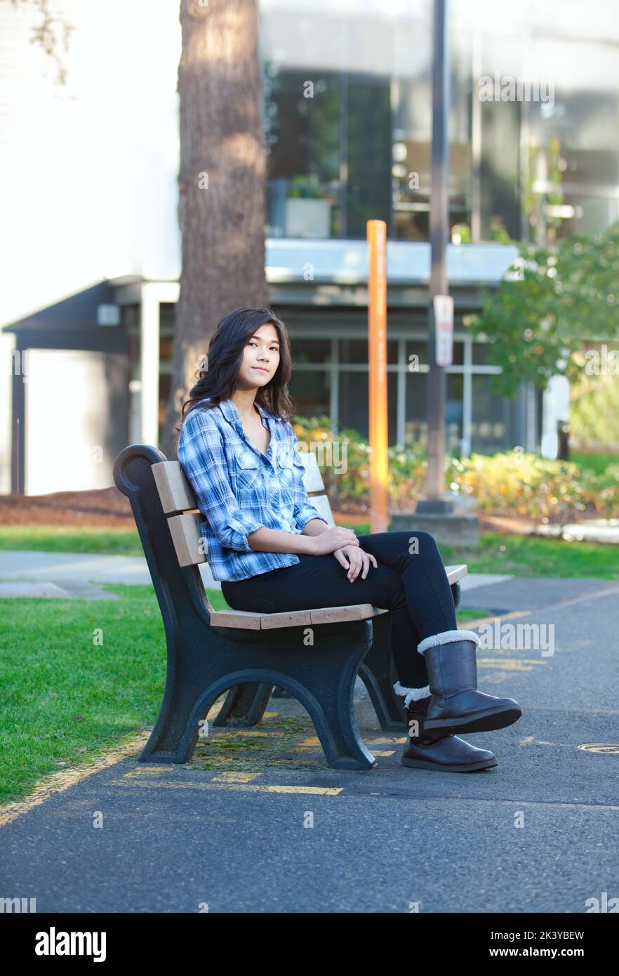 Jeune fille asiatique biracial assise sur un banc à l'extérieur du bâtiment de l'école, jambes croisées Banque D'Images
