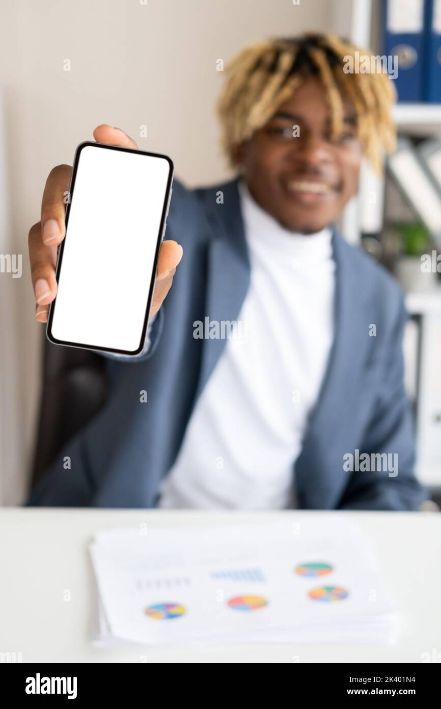 Communication d'entreprise. Joyeux homme noir. Application mobile. Beau gars souriant et défoqué montrant un smartphone écran vide, un bureau dans une pièce lumineuse Banque D'Images Communication d'entreprise. Joyeux homme noir. Application mobile. Beau gars souriant et défoqué montrant un smartphone écran vide, un bureau dans une pièce lumineuse Banque D'Images