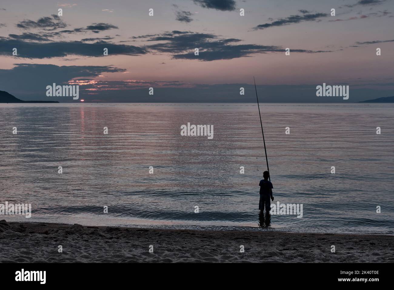Silhouette de garçon pêchant avec canne à pêche sur la rive sablonneuse du lac pendant le coucher du soleil. Banque D'Images