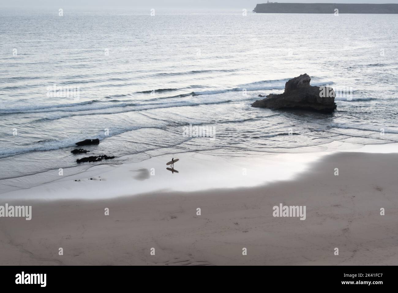 Sagres, Portugal - 30 mai 2022 : image monochrome d'une seule vague surfeuse à pied à la maison avec surf de la mer le long de la plage de sable au crépuscule à Sagres Portu Banque D'Images