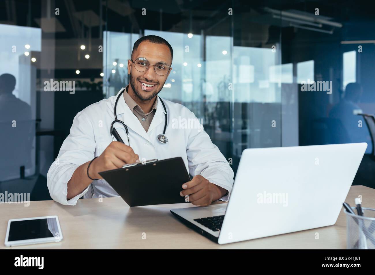 Portrait d'un médecin afro-américain, homme travaillant à l'intérieur d'un bureau de clinique moderne à table avec un ordinateur portable, médecin sous un manteau médical et stéthoscope souriant et regardant l'appareil photo. Banque D'Images