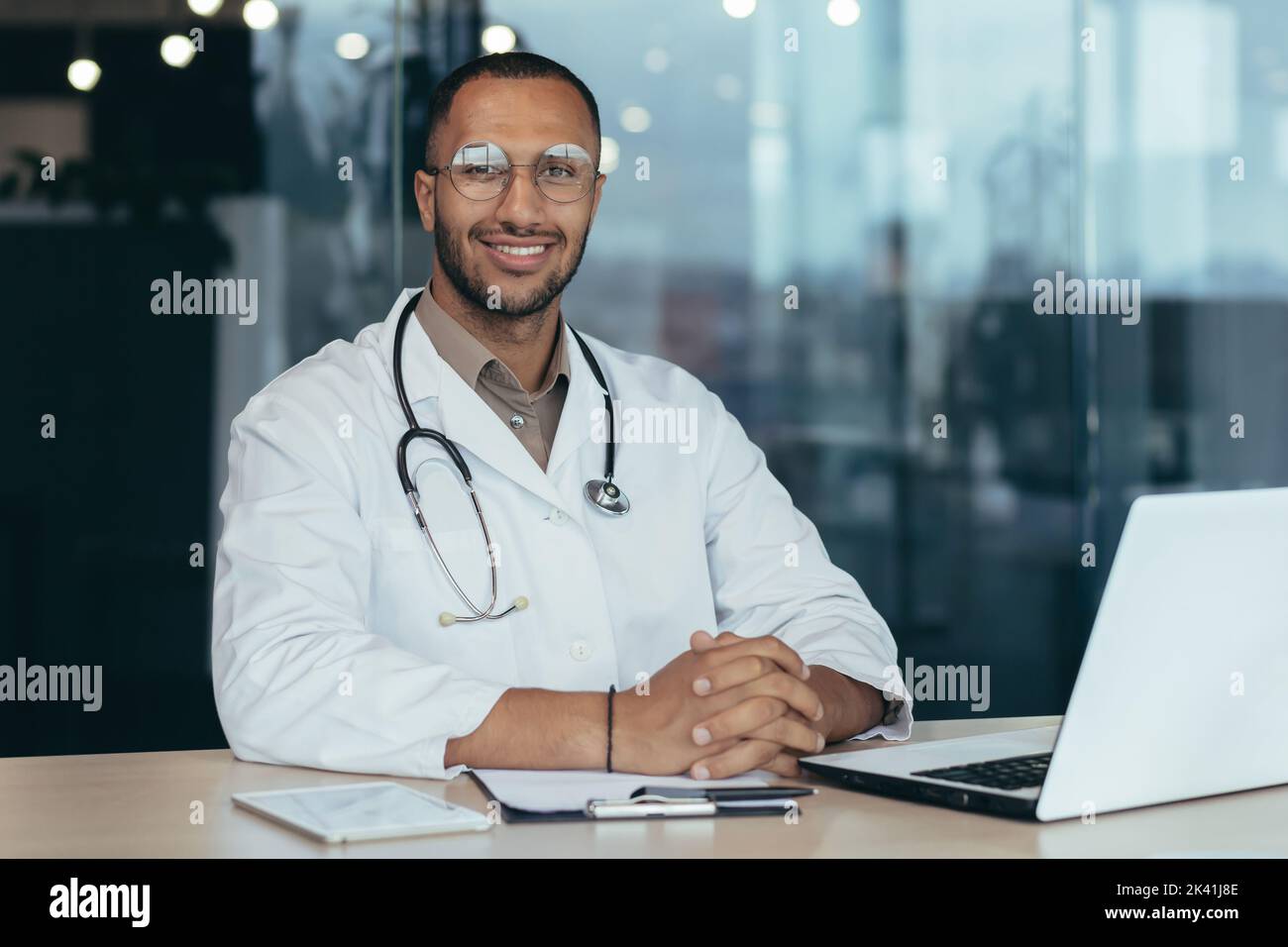 Portrait d'un médecin afro-américain, homme travaillant à l'intérieur d'un bureau de clinique moderne à table avec un ordinateur portable, médecin sous un manteau médical et stéthoscope souriant et regardant l'appareil photo. Banque D'Images