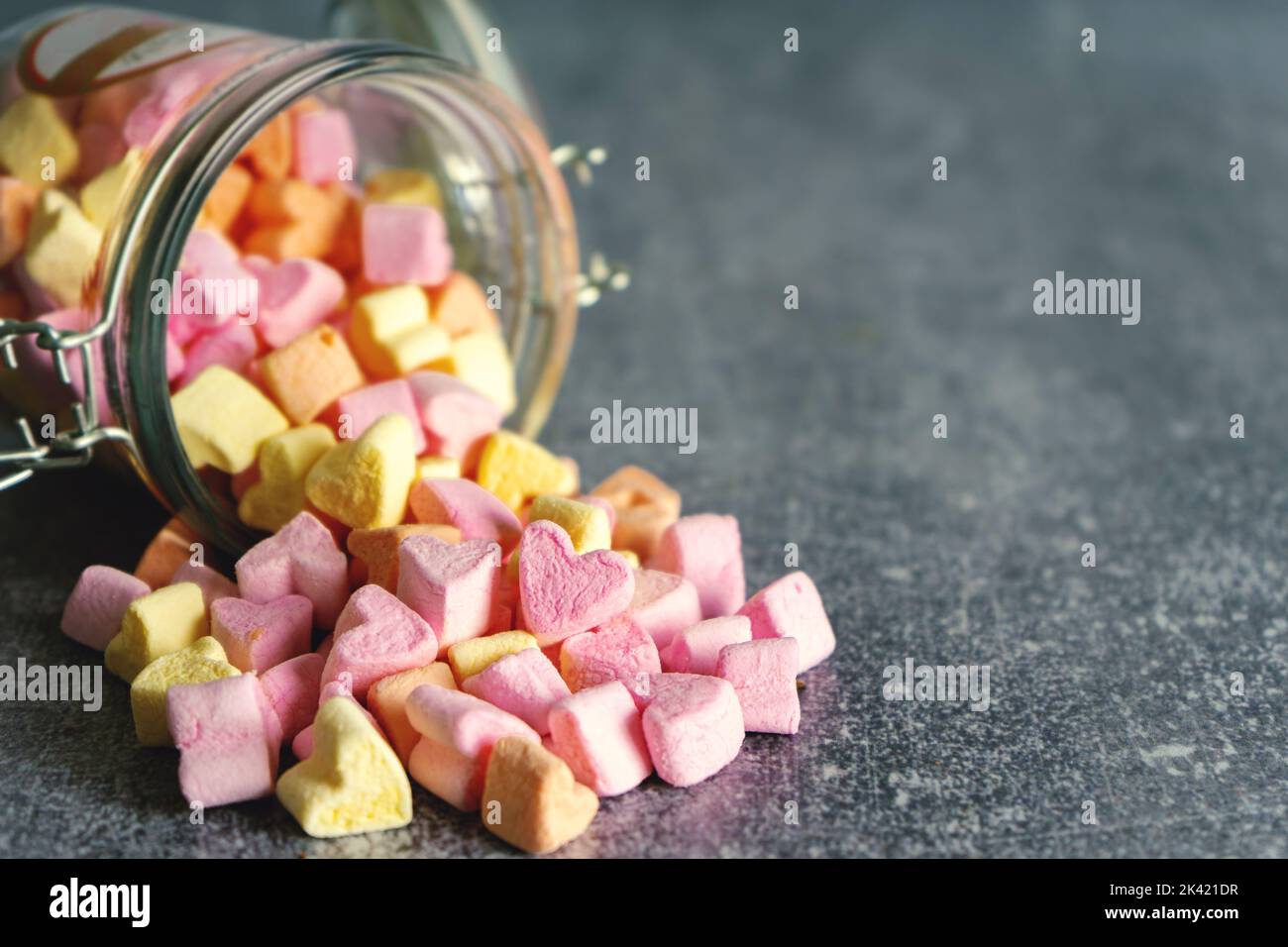 Tas de bonbons guimauves en forme de coeur rose et jaune, dispersés dans un pot en verre sur fond gris avec espace de copie Banque D'Images