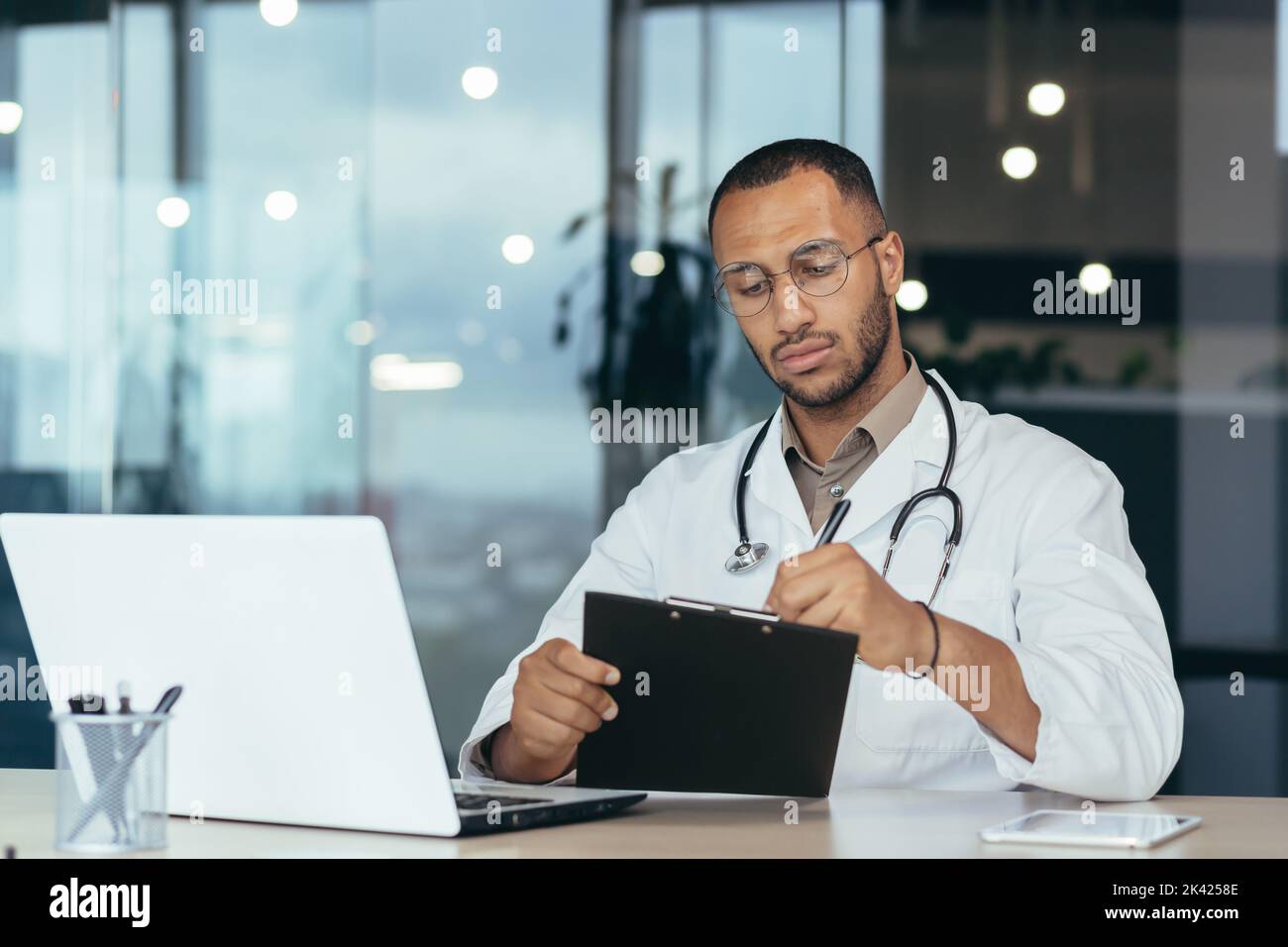 Médecin hispanique sérieux et concentré en blouse médicale et stéthoscope derrière la paperasserie, homme en lunettes travaillant à l'intérieur du bureau de la clinique à l'aide d'un ordinateur portable. Banque D'Images
