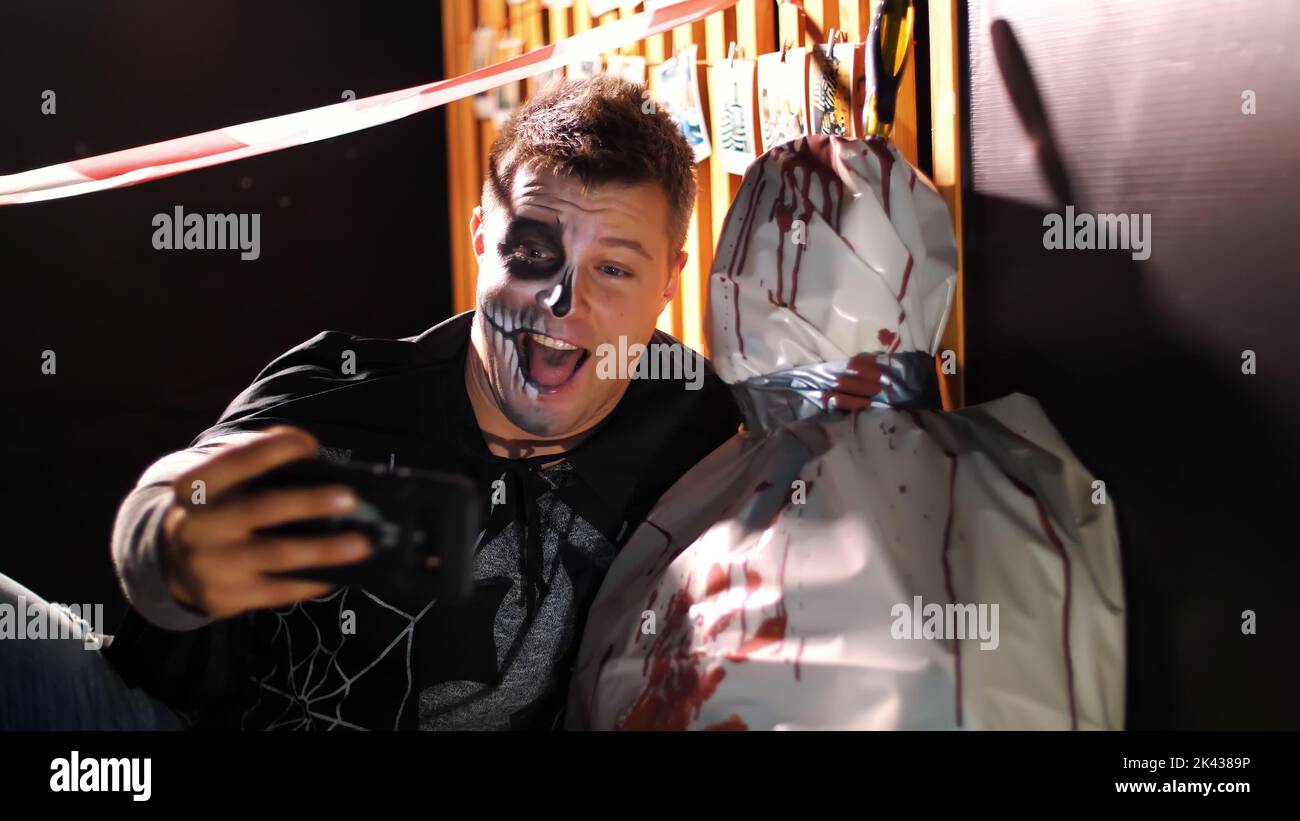 Fête d'Halloween, nuit, crépuscule, dans les rayons de lumière, un homme avec un maquillage terrible fait selfie avec un cadavre enveloppé d'un tissu d'huile, le cadavre est enduit de sang. Photo de haute qualité Banque D'Images