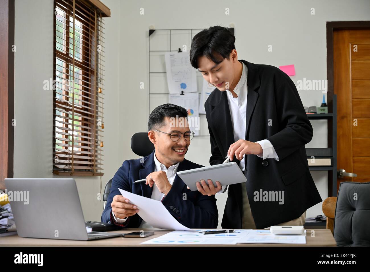 Homme d'affaires asiatique intelligent et prospère travaillant au bureau, en consultant son calendrier de réunions sur tablette avec son jeune assistant. Banque D'Images