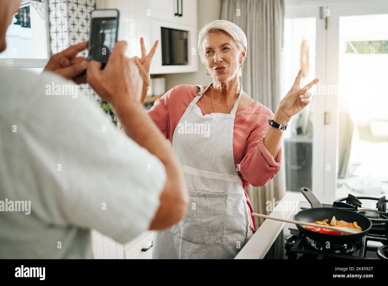 Un couple senior qui fait la cuisine, un smartphone qui prend le portrait en cuisine et une femme avec le signe de paix v pour les médias sociaux, la célébration de la retraite ou la santé Banque D'Images