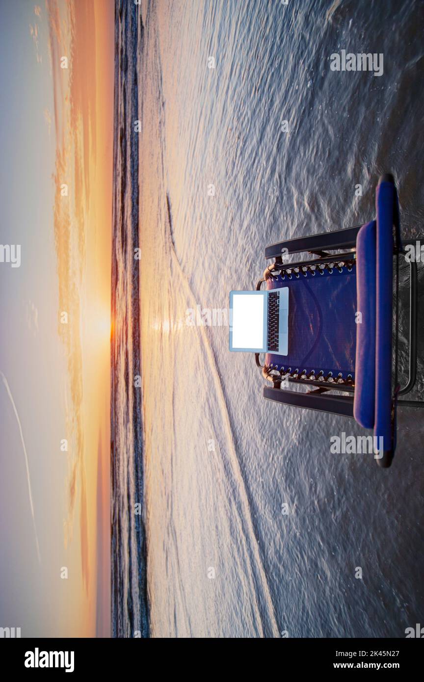 Ordinateur portable ouvert sur une chaise longue en vague peu profonde à la plage au coucher du soleil. Banque D'Images