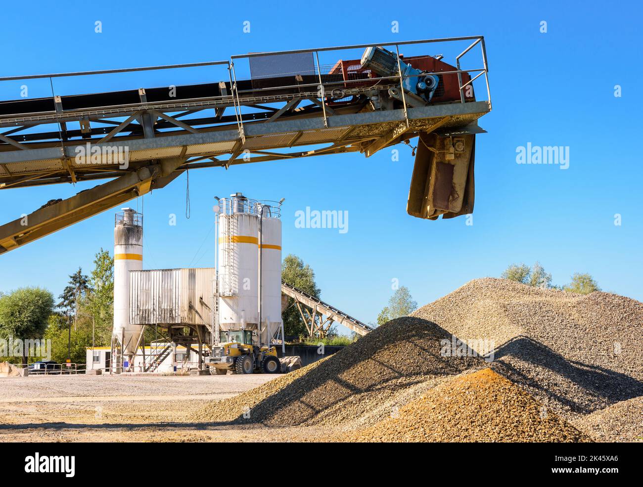 Un tapis roulant au-dessus de piles de gravier et une chargeuse sur pneus garés à côté d'un silo de sable dans une carrière par temps ensoleillé. Banque D'Images