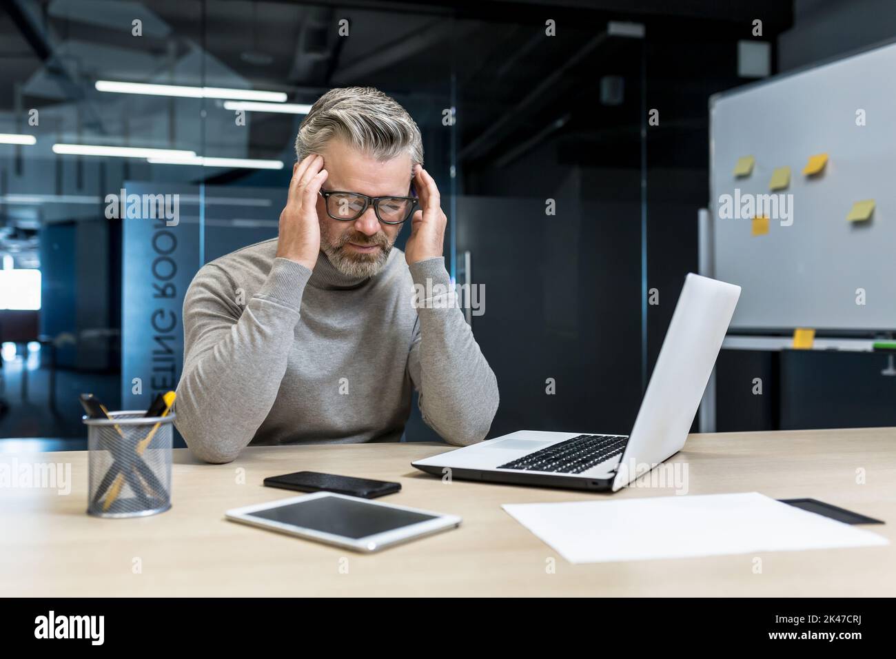 Maux de tête de tension au travail. Un jeune homme d'affaires aux cheveux gris élégant, en lunettes, est assis dans le bureau à un bureau avec un ordinateur portable et un téléphone, ressent un mal de tête, tient sa tête avec les mains. Banque D'Images