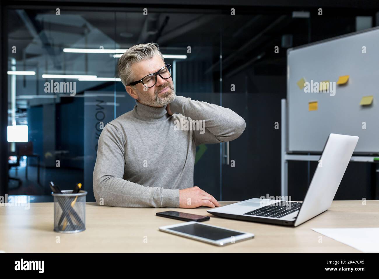 Un homme d'affaires fatigué et épuisé en lunettes est assis dans le bureau au bureau, tient son cou, moque son cou avec sa main. Il ressent de la douleur dans ses muscles. Banque D'Images