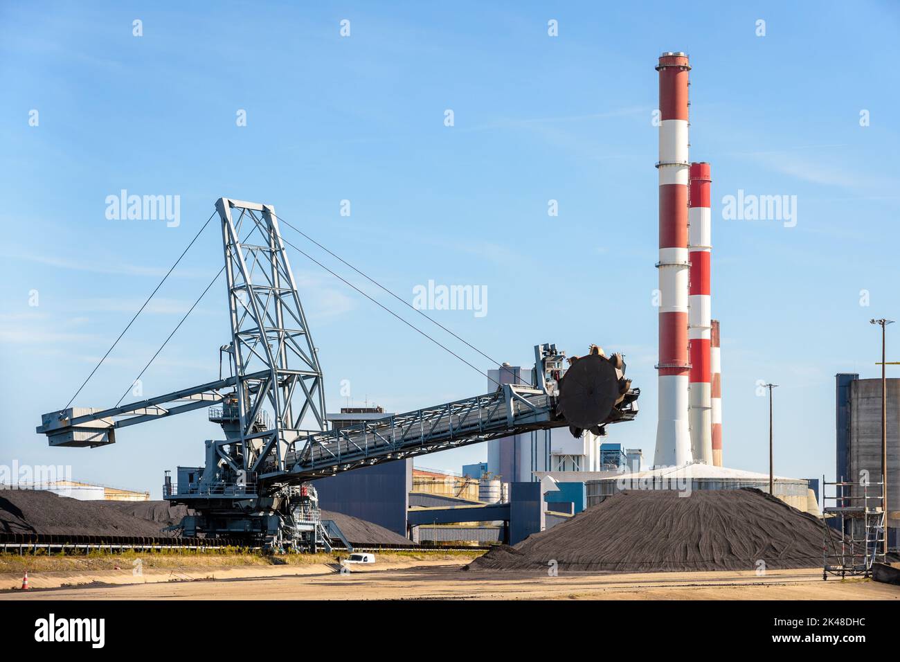 Un gerbeur-récupérateur à roulettes et des stocks de charbon dans une centrale thermique avec des cheminées rouges et blanches par une journée ensoleillée. Banque D'Images