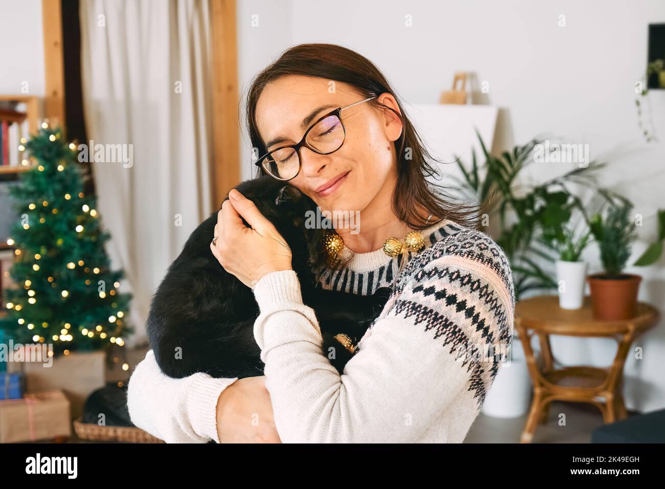 Jeune femme souriante embrassant chat domestique noir dans des lumières de Noël festives dans une pièce moderne avec des cadeaux et la décoration de noël. Joyeuses vacances d'hiver Banque D'Images