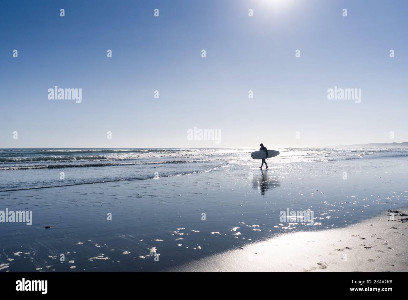 Homme rétroéclairé marchant le long de la plage avec une planche de surf sous son bras Banque D'Images